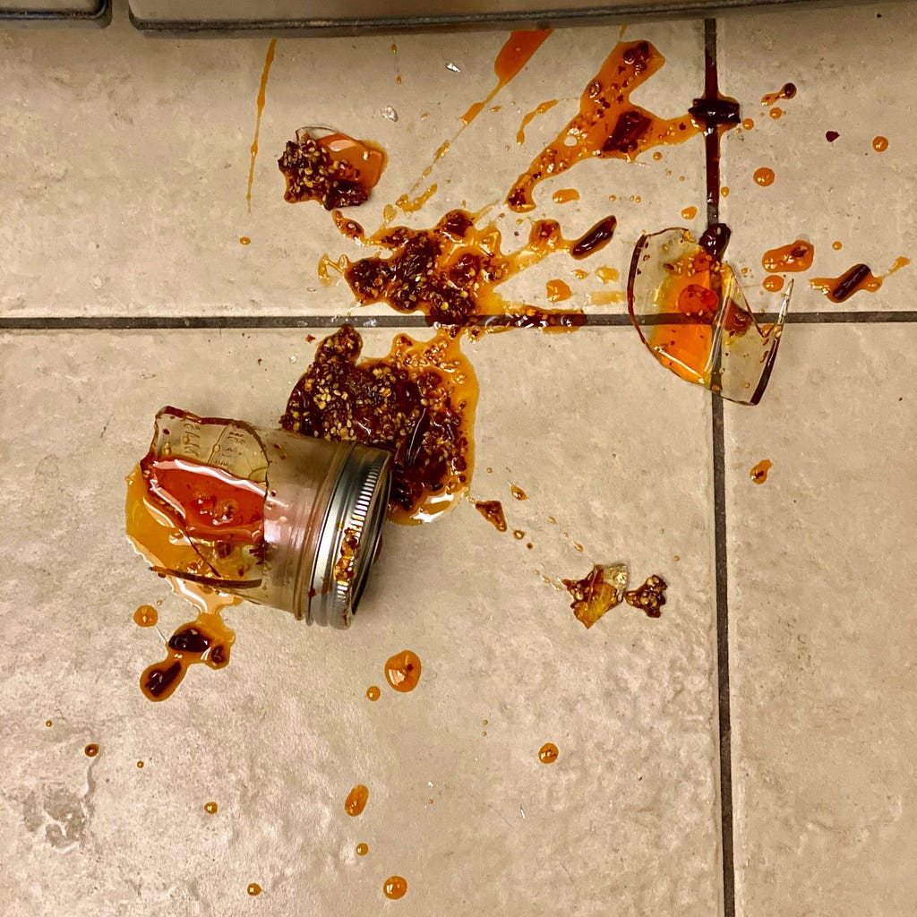 A shattered jar splatters a red chili oil across tiled floor.
