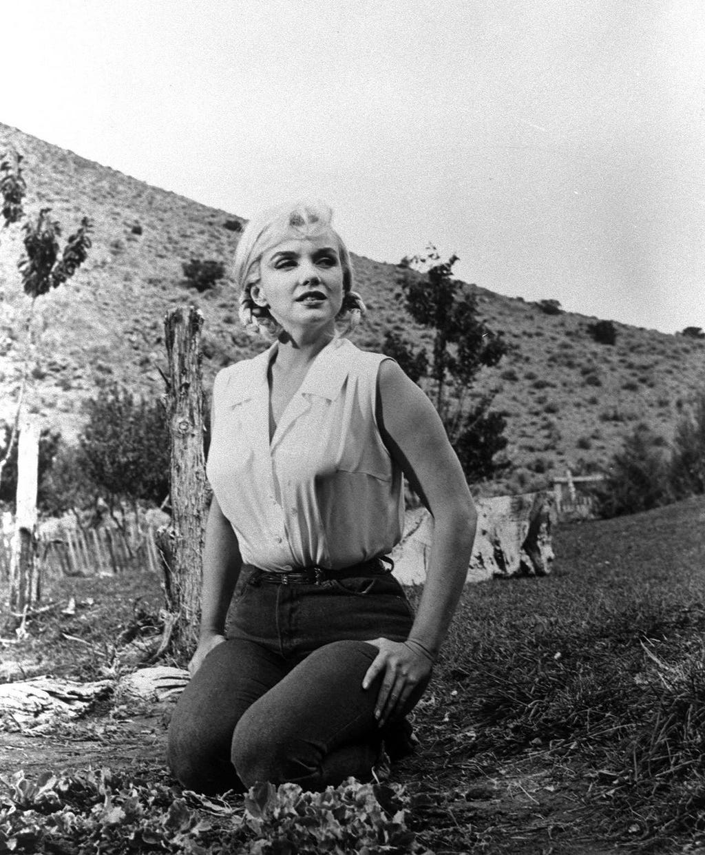 A black and white photo of Marilyn Monroe wearing a white tank top tucked into high-waisted jeans. She’s sitting on her knees