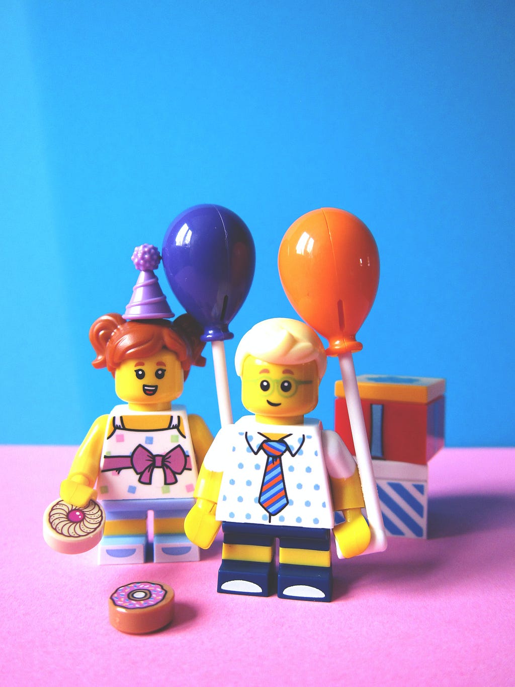 A Lego boy and girl in party attire hold balloons next to a birthday cake.