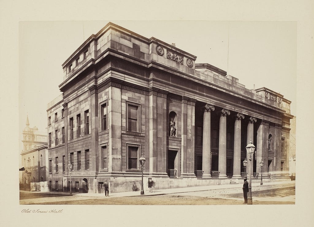 Manchester’s first Town Hall. Neo-classical design with a colonnade of four Ionic columns, and statuary. Four pedestrians.