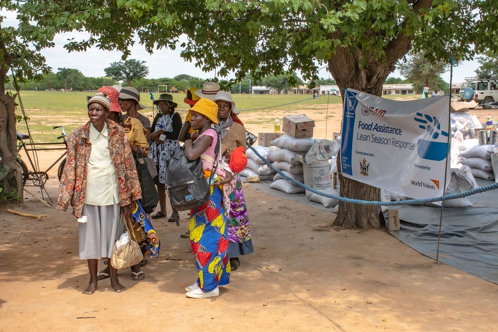 A group of women stand under a tree and next to sacks of food aid with a sign that says “USAID Food Assistance Lean Season Response.”