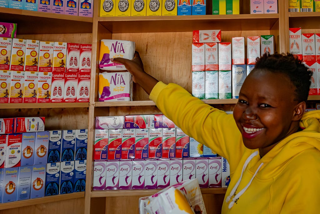 A smiling woman in a yellow sweatshirt stacks menstrual pads on a store shelf.