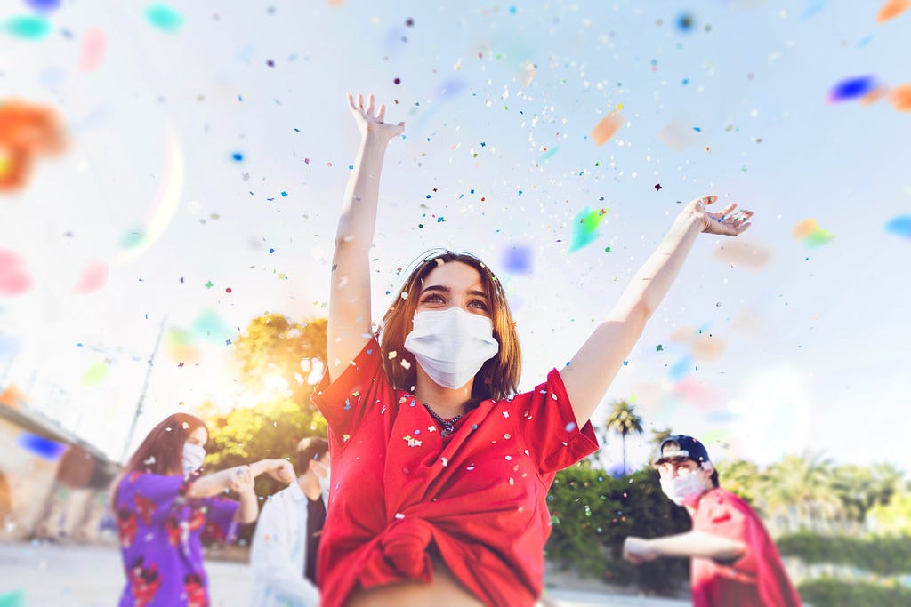 Woman wearing a face mask throws her arms up in celebration with her friends in the background and confetti in the air.