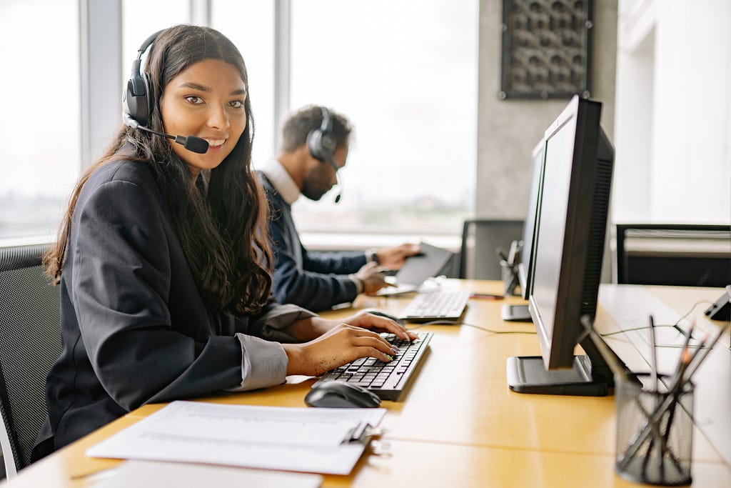 Picture of a young women, wearing a headset, at a computer, typing, with a man, also wearing a headset and typing, in the background.