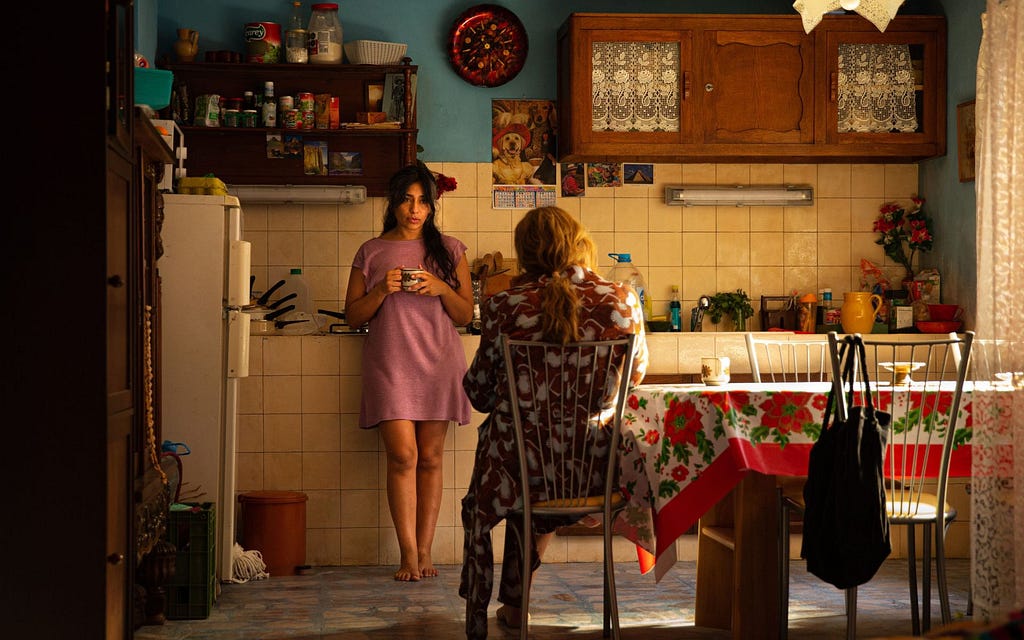 Shot of Adriana Paz as Epifanía, and Karla Sofía Gascón as Emilia Pérez. They are in a kitchen, Gascón sitting at a table with her back to the camera.