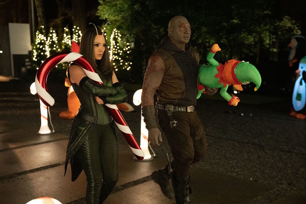 Mantis and Drax walking together. Mantis is carrying a giant candy cane while Drax carries a blow-up elf decoration.