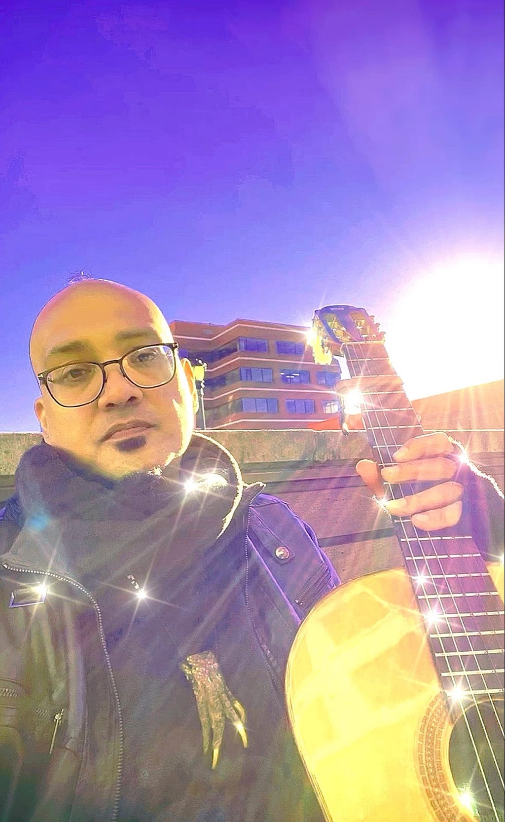 An image of the author, with a shaved head and glasses, holding a guitar in the air outdoors as the sun shines down from behind over the top of the buildings dotting the cityscape.