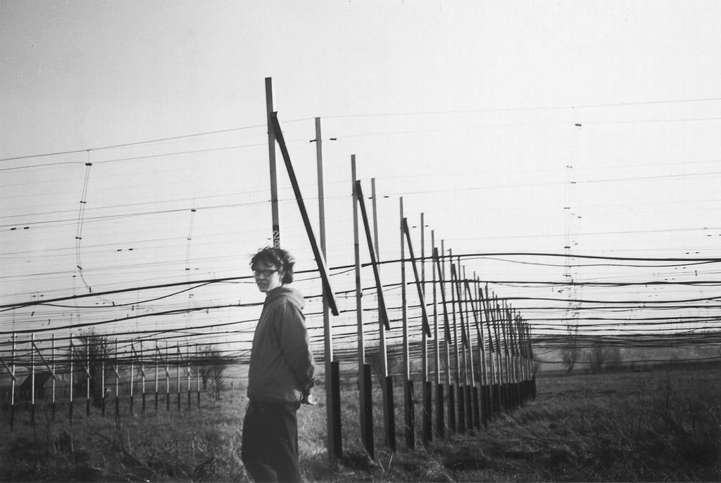 A black and white photo of a young white woman with short hair and glasses standing in front of a large series of poles that have been pounded into the ground in a large grid-like formation. Between each pole, numerous cables have been strung, creating what looks somewhat like a large floating net about three metres off the ground.