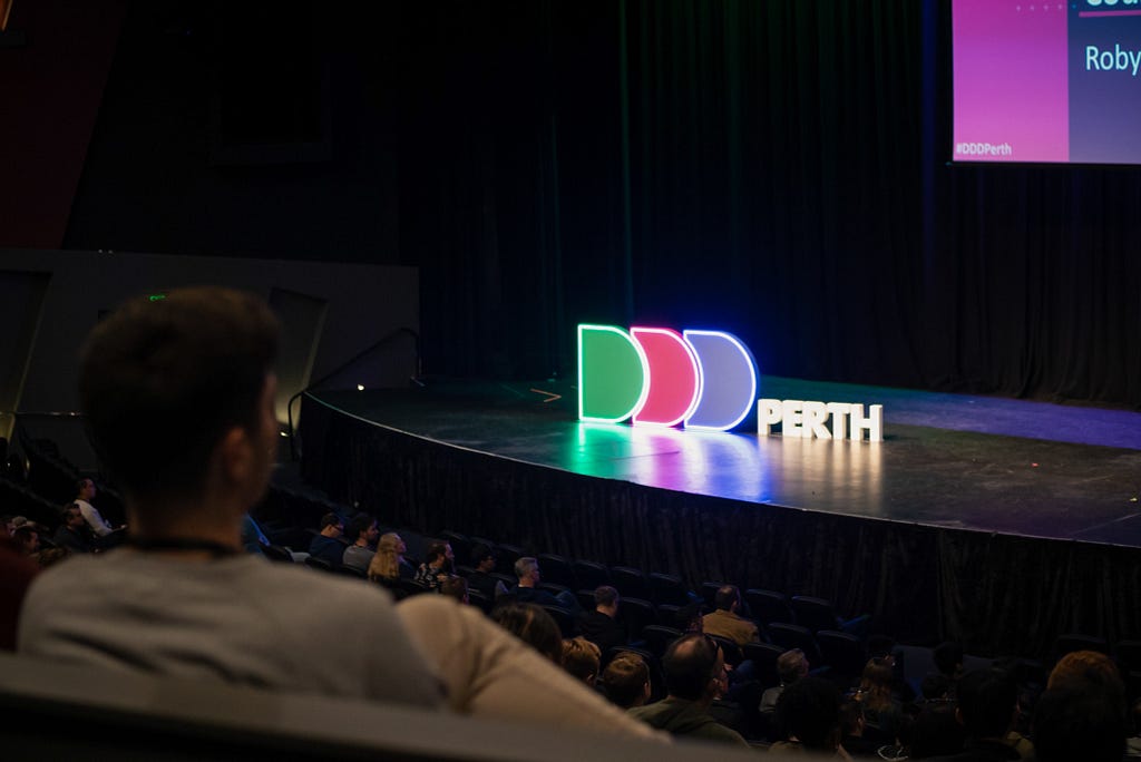 The DDD Perth neon sign on stage at PCEC. The photo is taken from a distance in a full theatre, and directly in front to the left of the camera is a person sitting with their back to the camera, facing the stage. They have their leg crossed.