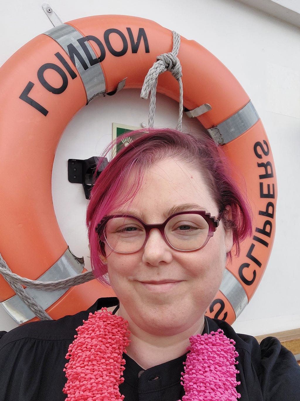 A picture of Claire with pink hair and a statement necklace, in front of a lifebuoy ring with “London Clipper” written on it