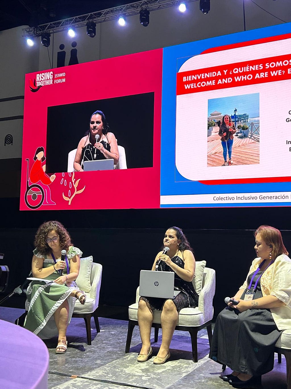 Tres mujeres posan sentadas frente a una pantalla en el escenario principal del Forum AWID frente a una audiencia. Dos de ellas sostienen un micrófono. una de ellas sostiene una laptop, preparándose para hablar.