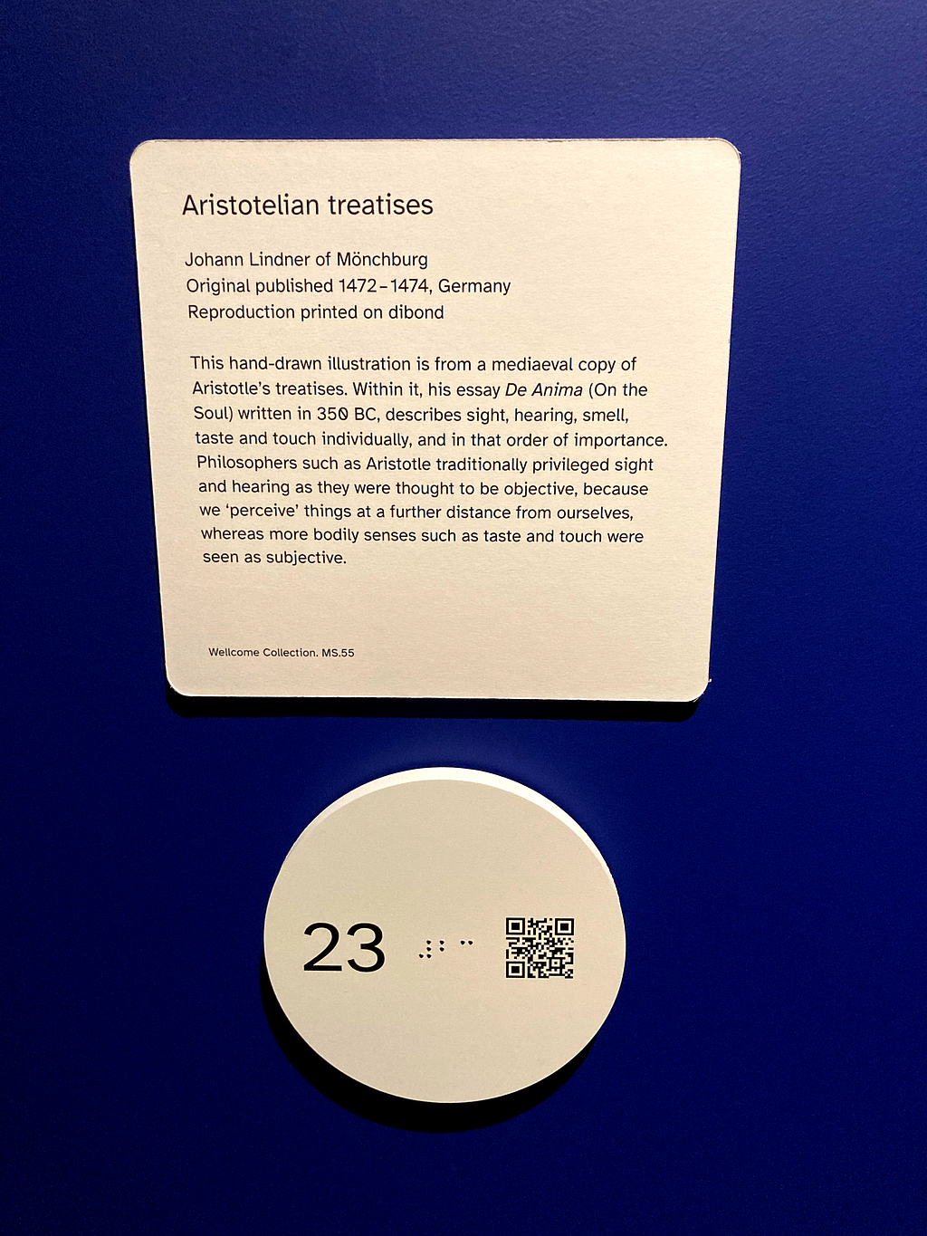 A photograph showing white signs in the gallery space on a blue wall. There is a rectangular sign with black text explaining the details of the exhibition item. Below this is a tactile sign that is round and raised, on it is a number refering to the stop in the guide, braile and a QR code that leads the to the digital exhibition guide when scanned