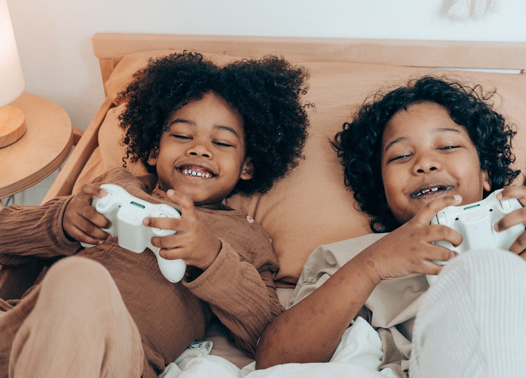 Two kids on a bed, smiling and holding video game controls with their hands.