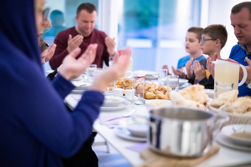 An Islamic family praying before iftar. There are adults and children raising their hands in prayer, with a selection of food ready to eat on the table.