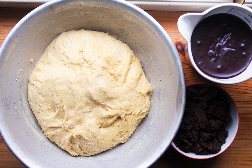 Chocolate babka ingredients viewed from above. A large bowl of dough is on the left, a jug of chocolate ganache and small bowl of chocolate streusel on the right.