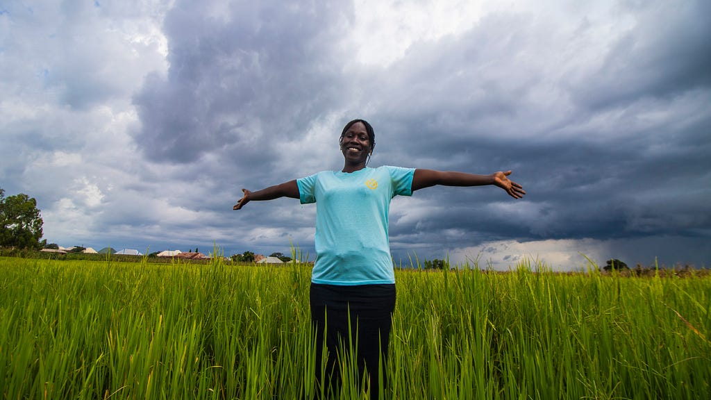 A smiling woman stands with arms outstretched in a rice field in Nigeria.