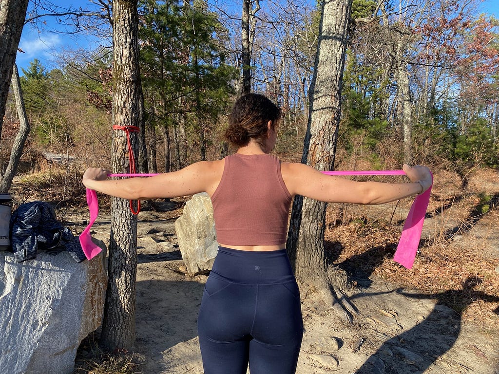 a woman performing resistance band exercises with a pink resistance band outdoors at a campsite