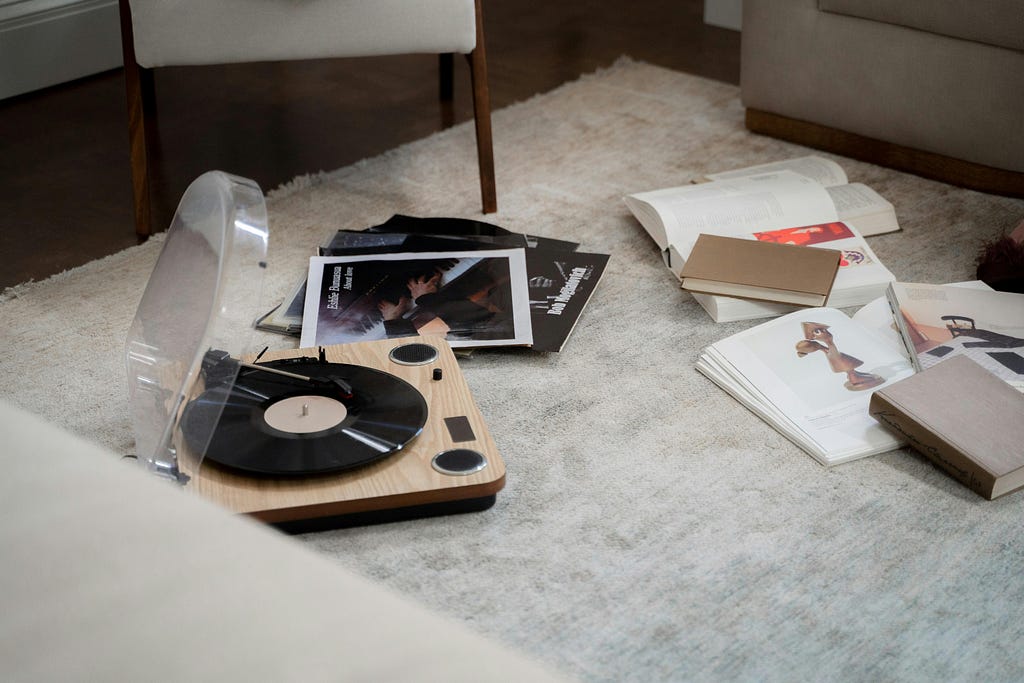 A record player sits on a soft rug on the floor with the lid open and a stack of vinyl records and books beside it. The corner of a white chair and couch frame the scene.