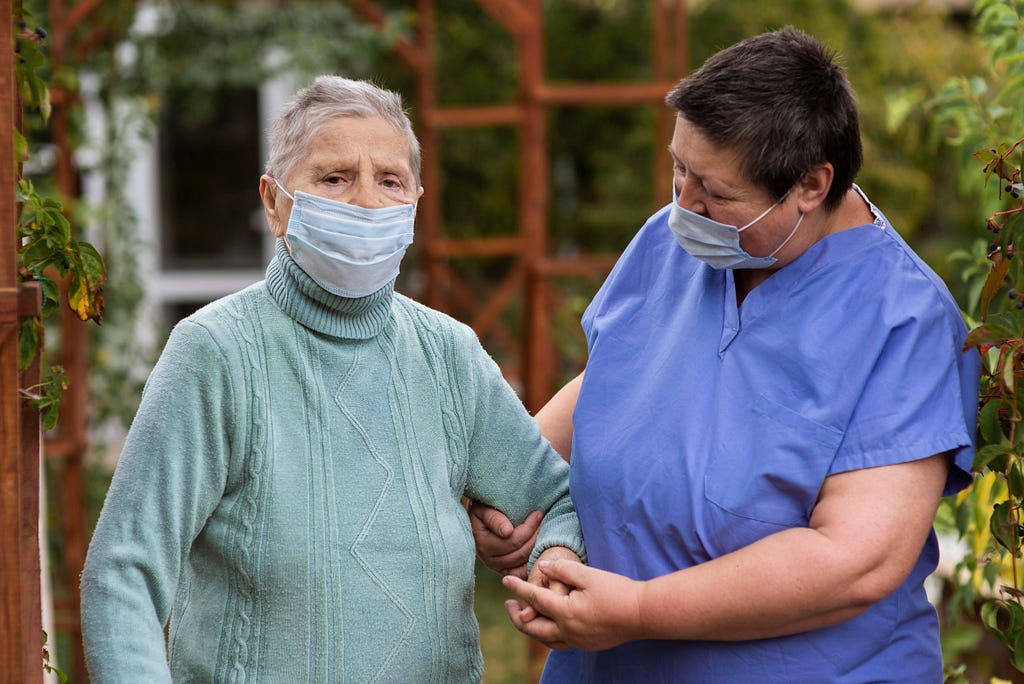 Enfermeira auxiliando idosa. Ambas de máscara.