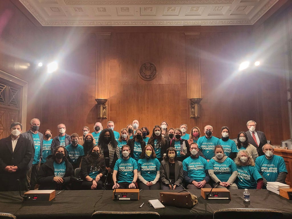 Members of the group Long Covid Moonshot and other advocates and researchers pose in a wood-paneled government room, wearing teal T-shirts advertising the need for moonshot-level funding.