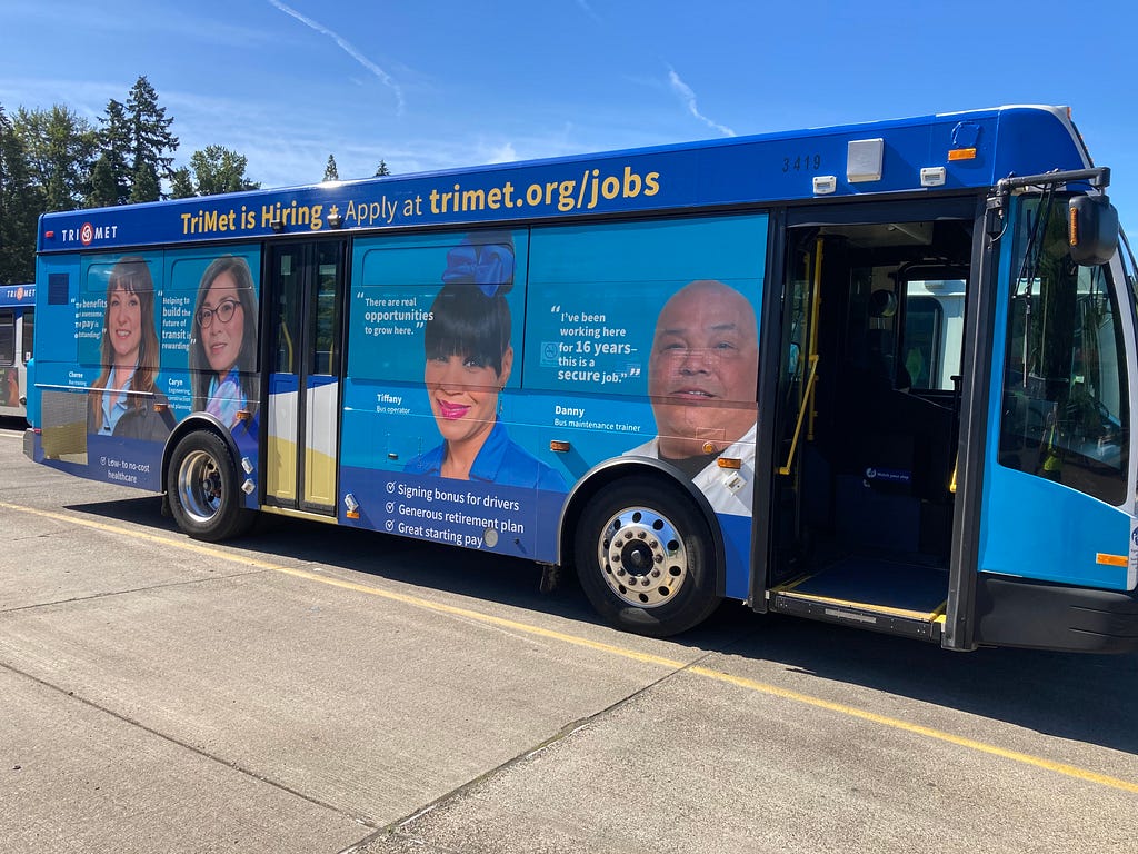 A Gillig 30 foot bus operated by TriMet wrapped with an advertisement about job opportunities at TriMet. The ad features four TriMet employees talking about how much they enjoy working at the transit agency. Cheree, a bus trainer says, “The benefits are awesome, the pay is outstanding.” Caryn, a planning engineer says, “Helping to build the future of transit is rewarding.”