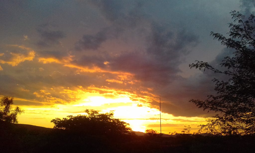 A black silhouette of the skyline with a mountain and trees as the sun is setting in a bright yellow sky and some cloud cover