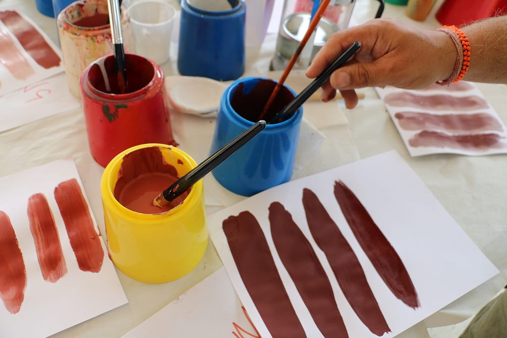 Paint pots with browns and orange paints. A hand reaches for one of several brushes in the pots over paper lines of colour.