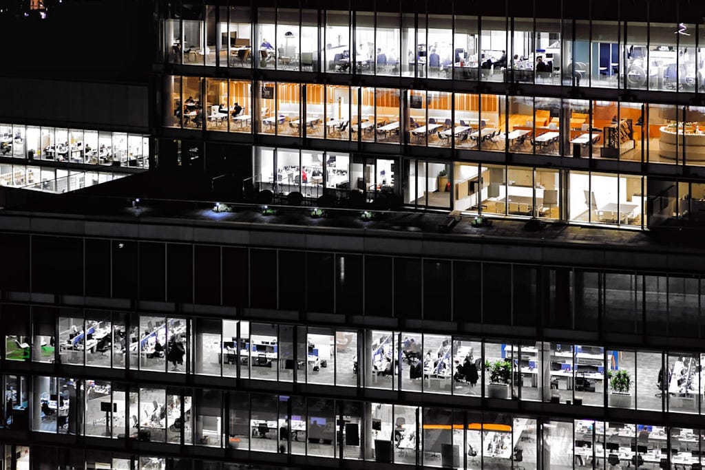 Looking into six floors of a glass commercial office tower at night we see sundry office furniture lined in rows inside. The top three floors are lit, the fourth from the top is dark and the bottom two also are lit.