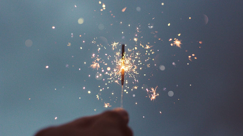 A hand holding a stick giving off little sparks (a firework sparkler) at night