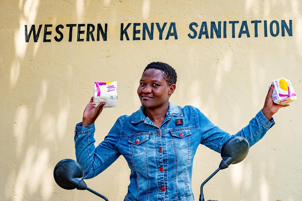 A smiling woman on a motorbike holds up menstrual pads in front of a wall with painted text that says: “Western Kenya Sanitation.”