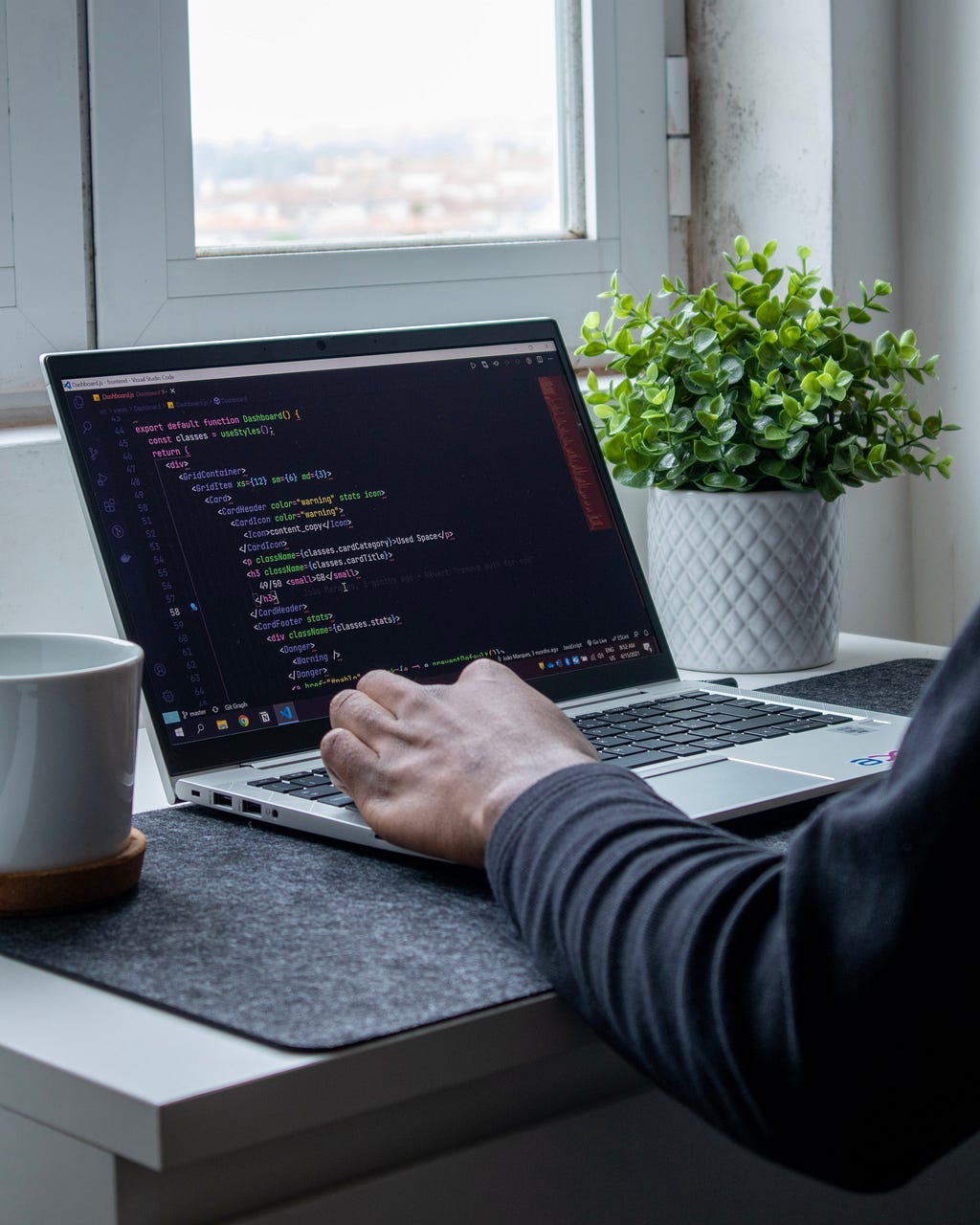 Lines of code showing on a screen of a MacBook Pro. A mug is on the left and a small plant on the right of the computer.