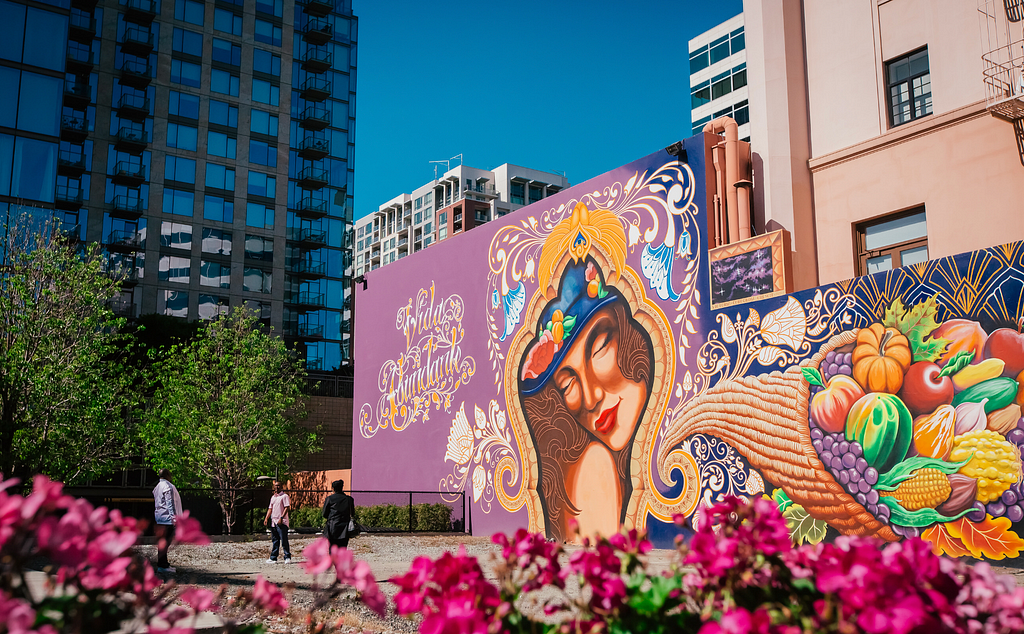 Photograph of an urban plaza with a large, colorful mural depicting a woman and a cornucopia of fruit.