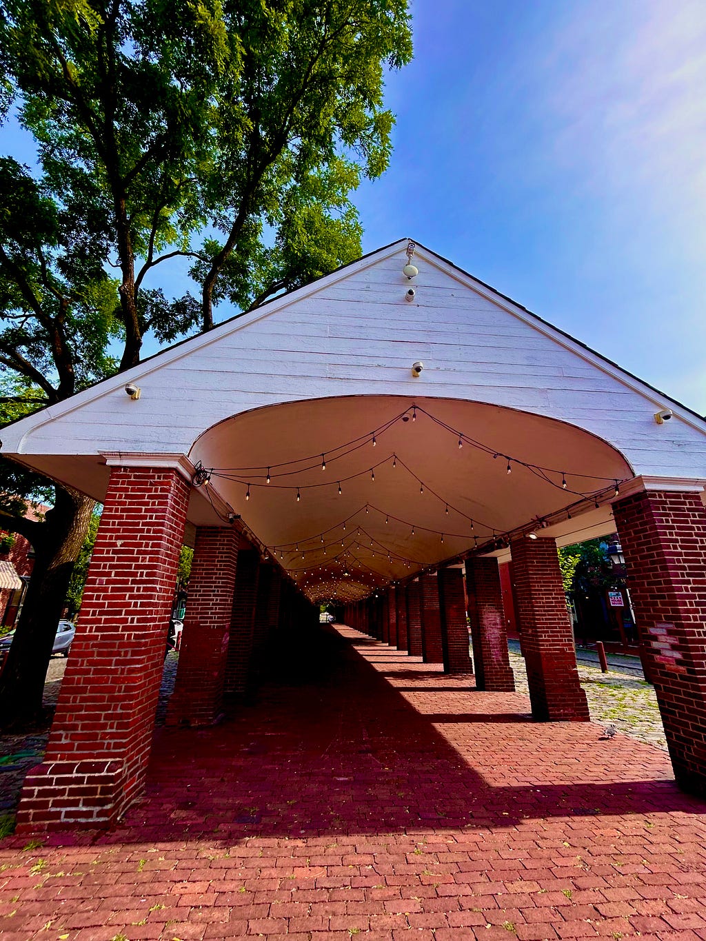 A covered walkway, with a triangular white roof supported by red brick columns during daylight.