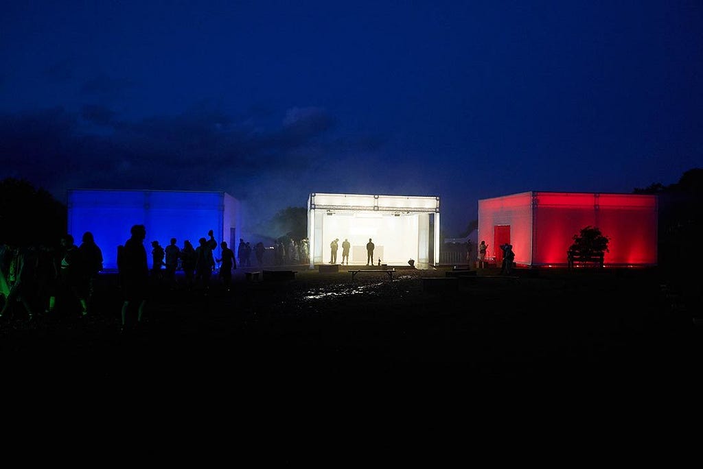 Es ist Nacht. Nebeneinander stehen 3 große Leuchtkästen in blau, weiß und rot.