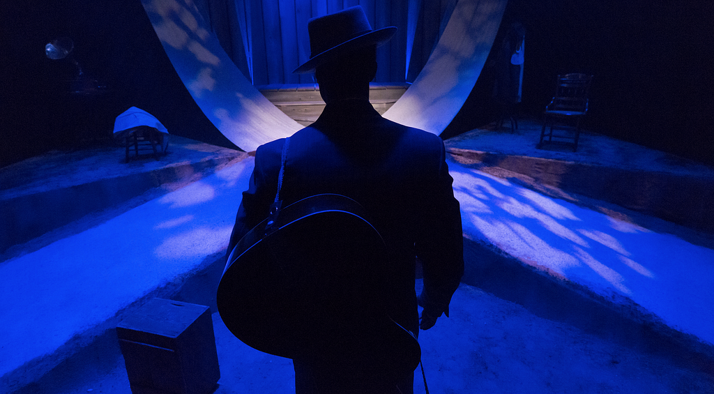 A man in a suit and hat with a guitar slung over his back stands with his back to the camera facing a blue-lit set