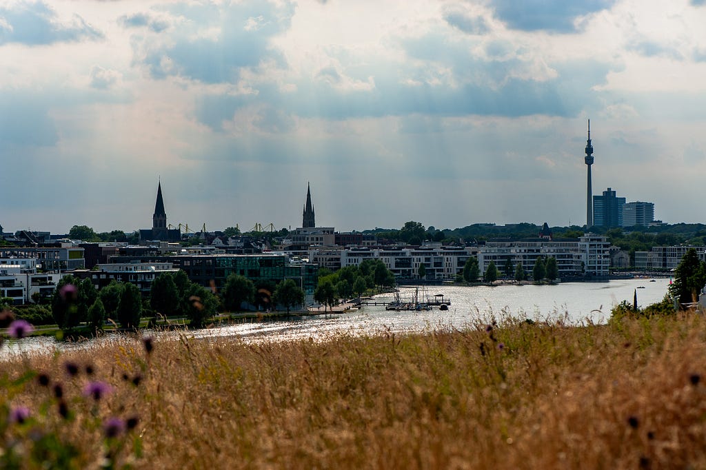 Looking west over the lake, the city’s skyline includes industrial reminders, churches, a tv tower, and the country’s largest soccer stadium. Dortmund, Germany, June 30, 2023.