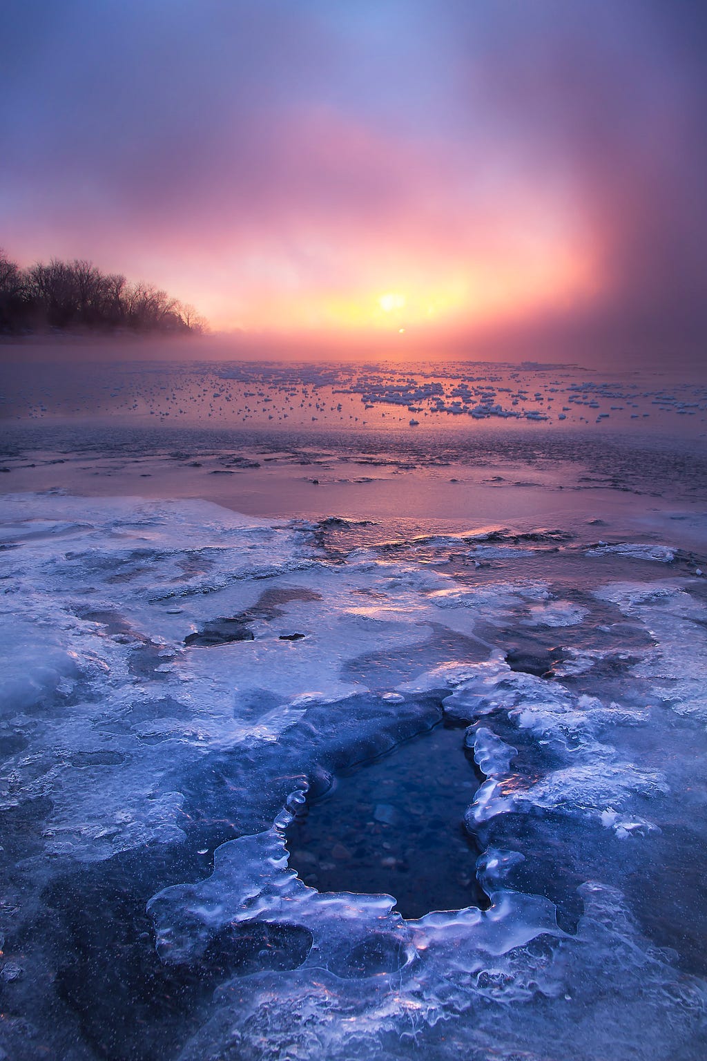 Sun rises through a pink and purple fog, reflected in the ice and water of a river.