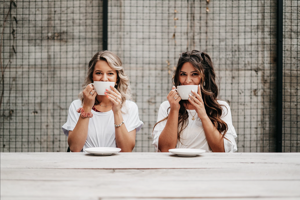 Two White women sitting next to each other wearing the same outfit, holding identical coffee mugs in front of their faces, facing the camera