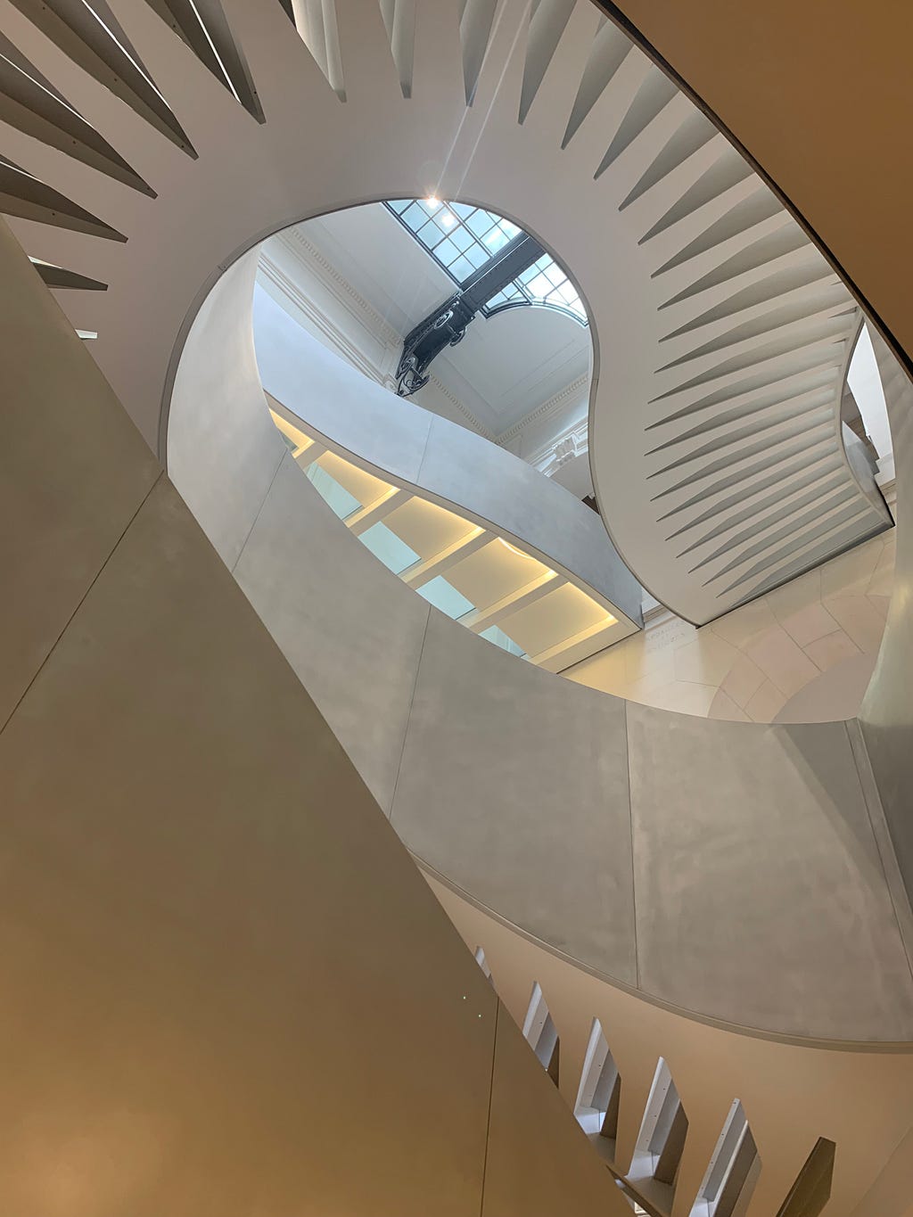 Low-angle view of a modern spiral staircase inside the BNF Richelieu. White steps emerge from a hollow center, surrounded by beige walls and cut-out balustrades. Above, a vaulted ceiling with windows lets in natural light.