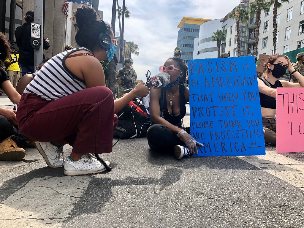 Reporter Emily Elena Dugdale holds a mic up to a person sitting on a street during a protest. The person protesting is holding a blue sign that reads “Racism is so American that when you protest it, people think you are protesting America.”