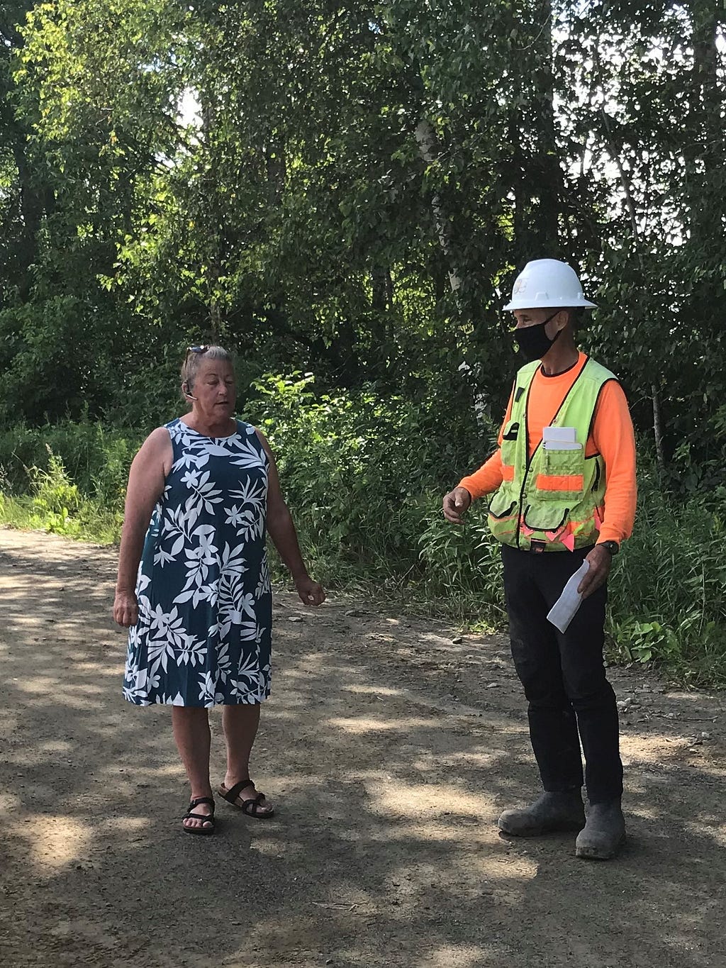 A woman in a dress stands next to a man in hardhat and safety vest outdoors.