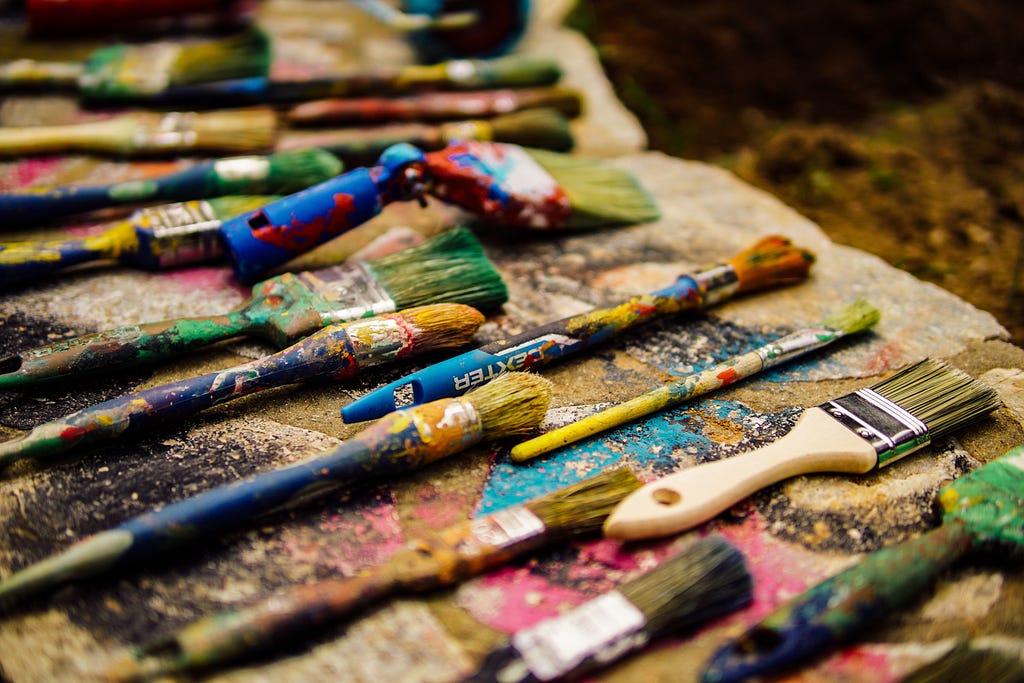 Many different sized paint brushes with messy paint. Brushes are laid out neatly in a row on a wooden slab.