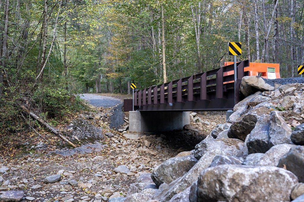 a metal bridge spans a low area along a road through the woods