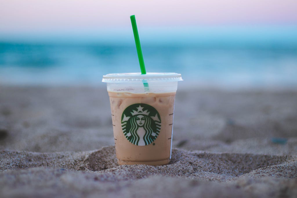 Starbucks iced coffee on a beach with blue ocean and pink sunset in the background.