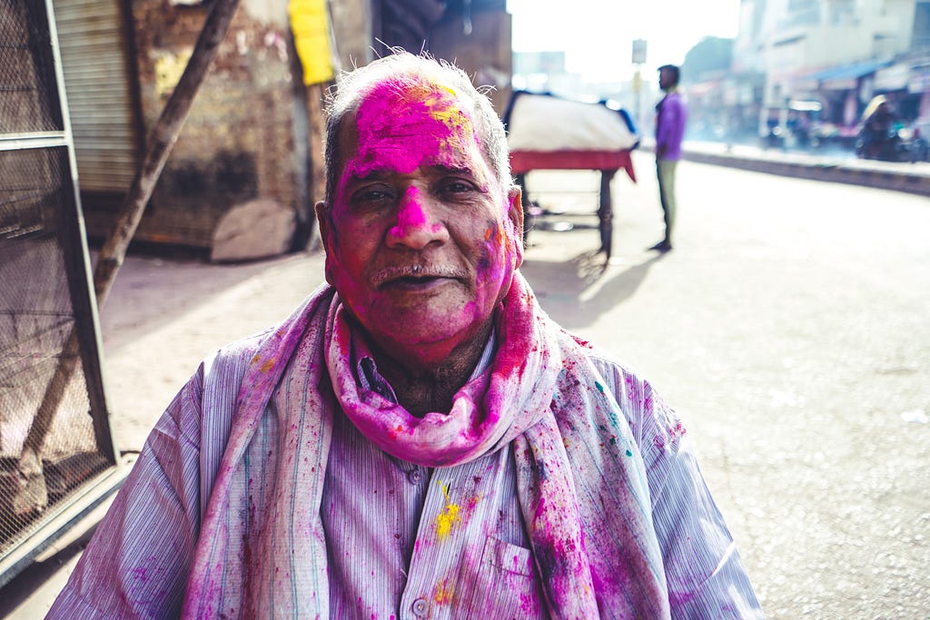 Street-photograph of an Indian man wearing a white shirt and who has strong purple color on his head and shirt with some yellow spots.