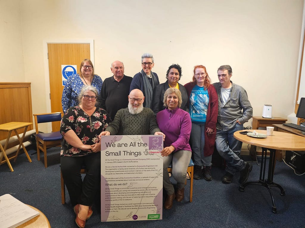 A diverse group of individuals standing and sitting in front of a colourful poster from All The Small Things.