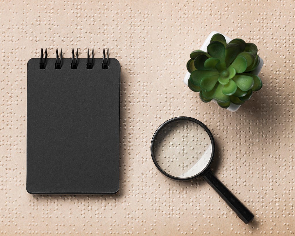 A notebook, a magnifying glass and a succulent on top of a Braille textured sheet. (Freepik)