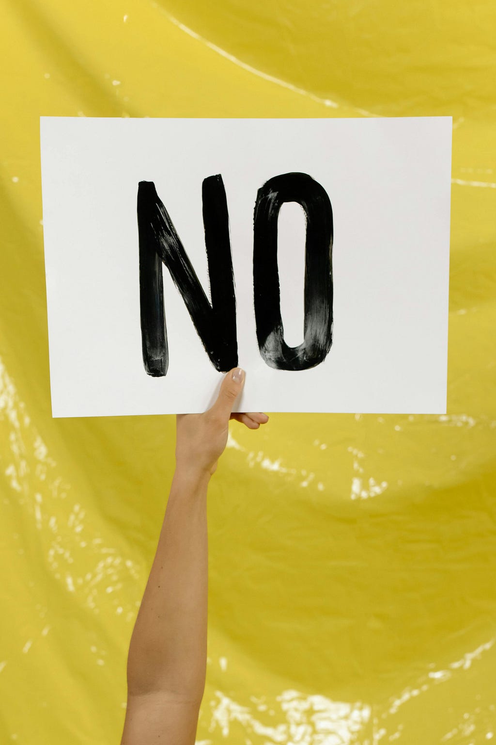 Human holding a banner saying ‘No’ against a yellow background. Source: Pexels.