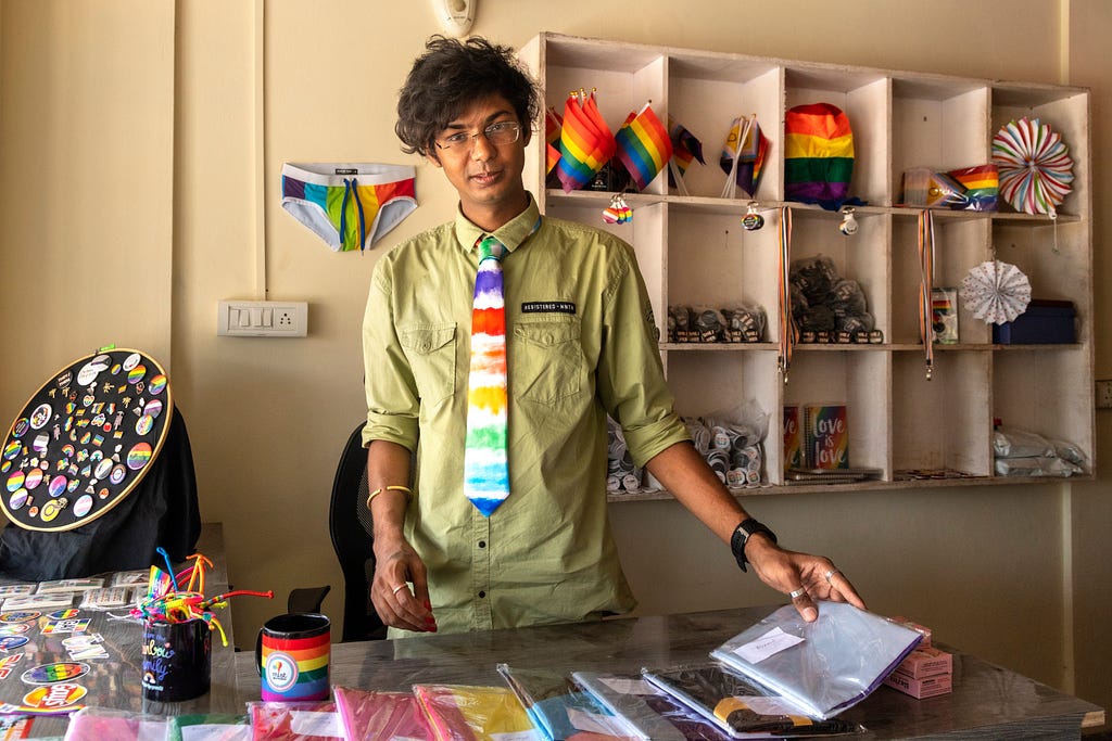 A man wearing a rainbow tie stands behind a store desk. Behind him the walls are covered in rainbow merchandise.