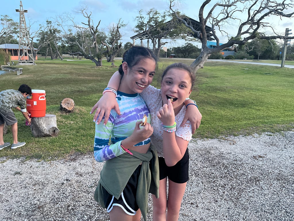 two campers hug and eat smores in front of the pavilion at camp